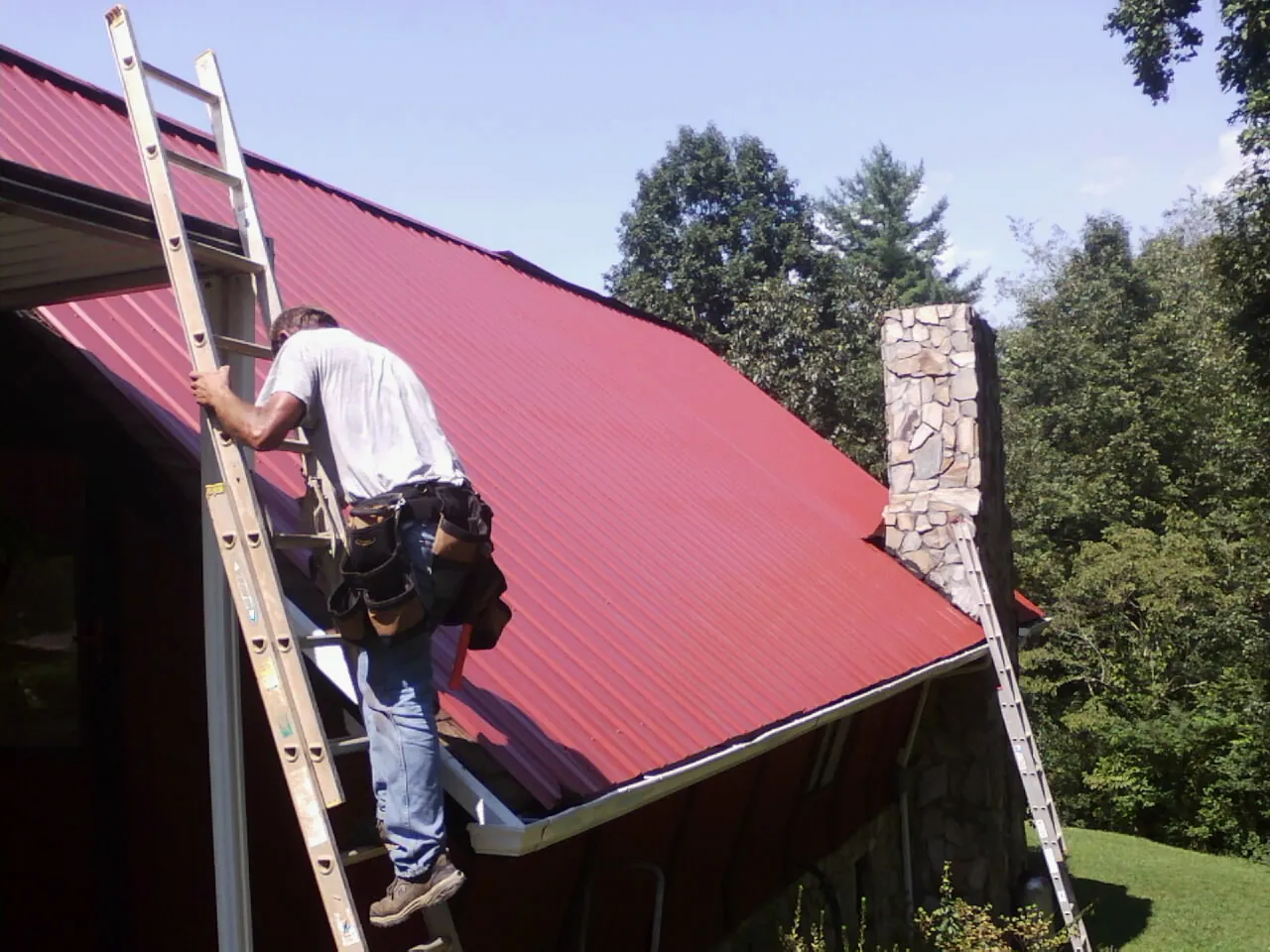 Roofer on a ladder beside a red metal roof and stone chimney — Soffit & Fascia Repair by Roofing Brothers in Meigs